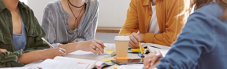 Vier menschen sitzen an einem Tisch, auf dem Tisch befindet sich Kaffee im Pappbecher , Unterlagen und Schreibmaterial.