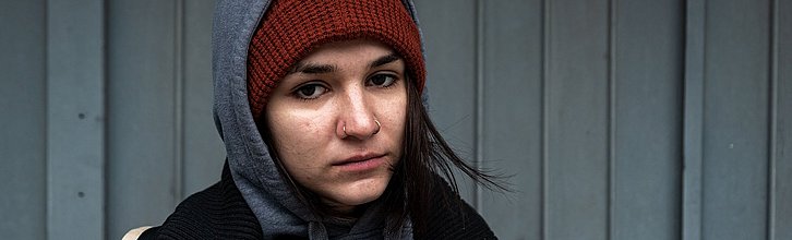 Frau mit schulterlangen dunkelbraunen Haaren, guckt frontal in die Kamera, hat lustlosen Blick, trägt Jacke, Mütze und Kapuzenpulli